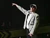 Macklemore i grå dress og caps peker mot venstre på en mørklagt scene.