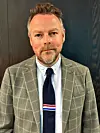 Mann med skjegg i rutete jakke og mørkt slips foran en mørk trevegg.