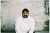 Aon Raza Naqvi sitter foran en slitt betongvegg iført hvit genser, caps og kjeder.