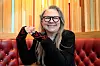 Mari Boine med briller smiler og gjør en håndbevegelse foran trepanel og rød skinnsofa.