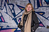 Kvinne i kåpe og skjerf foran en graffitivegg.
