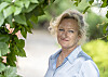 Trude Teige smiler mot kameraet i en lyseblå skjorte, innrammet av grønne blader.
