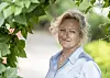 Trude Teige smiler mot kameraet i en lyseblå skjorte, innrammet av grønne blader.