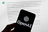 En mobiltelefon med OpenAI-logo foran en dataskjerm med tekst fra ChatGPT.