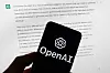 En mobiltelefon med OpenAI-logo foran en dataskjerm med tekst fra ChatGPT.