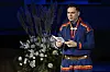 Sigbjørn Skåden holder et dokument på scenen ved siden av en stor blomsterdekorasjon.