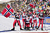 Ungdommer i skidrakter står samlet på snøen foran en stor folkemengde og norske flagg.