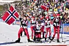 Ungdommer i skidrakter står samlet på snøen foran en stor folkemengde og norske flagg.