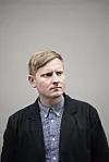 Blond mann i mørk jakke og blåmønstret skjorte foran en grå bakgrunn.